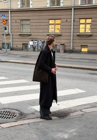 Duga crna vunena kapa širokog kroja. Stoji na pješačkom prijelazu, u kombinaciji s tamnim hlačama i crnim cipelama. U pozadini ulica i zgrada.