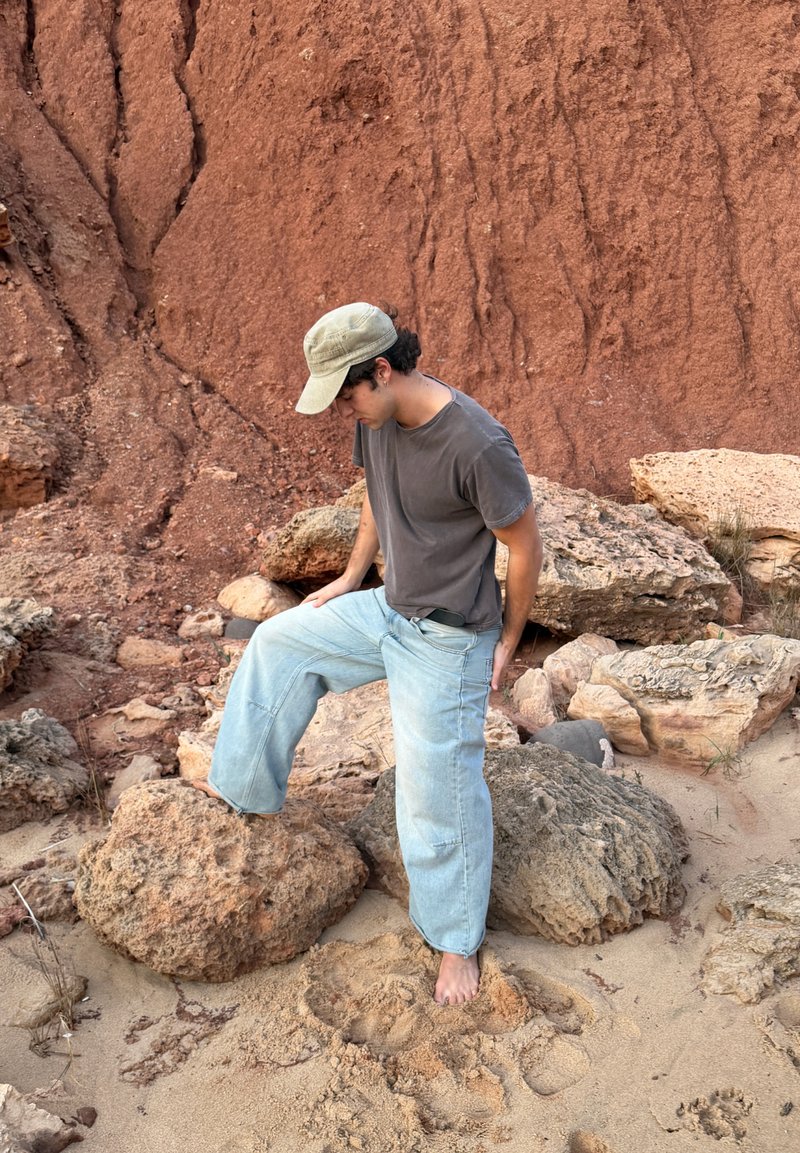 Jeune homme portant une casquette et une chemise grise, debout pieds nus sur un sol sablonneux, reposant un pied sur un gros rocher près d'une falaise rocheuse rouge.