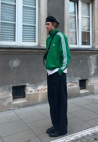 Zielona kurtka treningowa Adidas z białymi paskami, czarne spodnie i czarne buty. Kurtka ma zamek błyskawiczny i luźny krój. Stoi na zewnątrz.