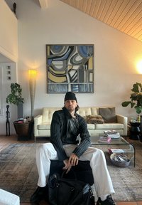 Sweter dziany w czarne paski, białe spodnie, czarna torba. Nowoczesny salon z beżową sofą, abstrakcyjną sztuką na ścianie, szklanym stolikiem kawowym i roślinami.