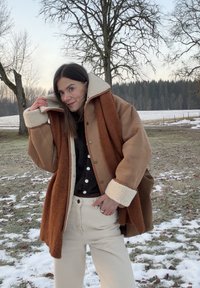 Femme portant une veste en peau retournée marron et une écharpe couleur rouille ajustant son col, debout sur un sol partiellement enneigé avec des arbres dénudés et une forêt en arrière-plan.