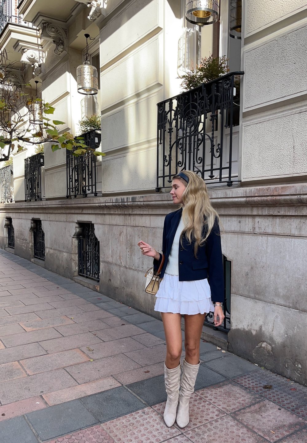 Ναυτικό σακάκι πάνω από λευκή μίνι φούστα με ρίγες, συνδυασμένο με μπεζ μπότες μέχρι το γόνατο και μια μικρή μπεζ τσάντα. Vintage-style δρόμος με επιχρυσωμένες ράβδους.