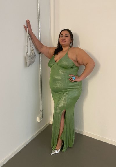 Vestido bodycon verde, brillante, con una abertura frontal, combinado con tacones metálicos plateados y un pequeño bolso plateado. Anillo azul como acento en la mano.