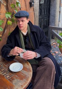 Person trägt einen dunklen Wollmantel und eine Schiebermütze, hält eine weiße Tasse mit silbernen Akzenten und sitzt an einem Holztisch mit Blättern und einem Teller in der Nähe.