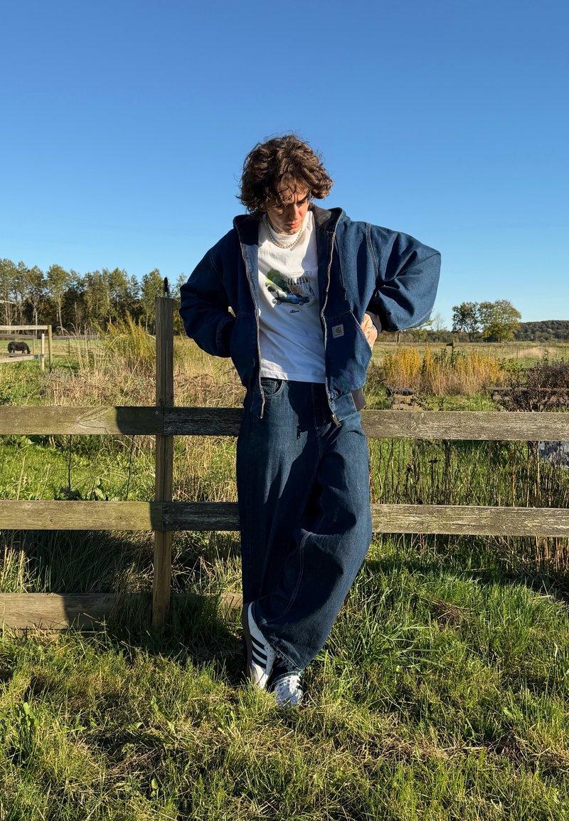 Jeansjacke mit Kapuze, hellblaues Shirt mit Grafik, locker sitzende dunkelblaue Jeans, weiße Sneaker, Hintergrund mit grünem Gras und Holzzaun.