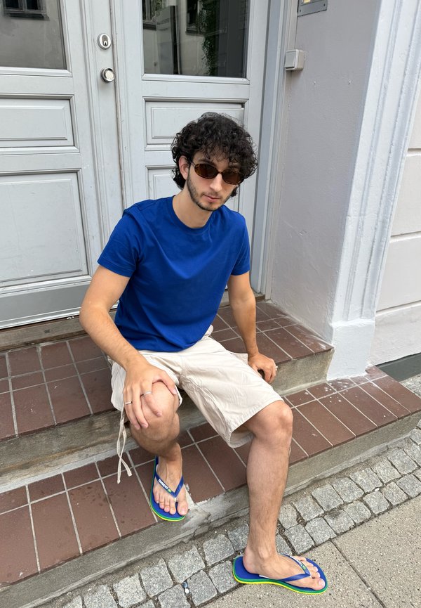Jeune homme aux cheveux bouclés et lunettes de soleil assis sur des marches extérieures, portant une chemise bleue, un short beige et des tongs bleues.