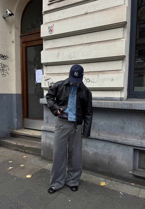 Juodas odinis švarkas ant džinsinio marškinėlių, pilkos smailinės plačios kelnės ir tamsiai mėlyna kepurė. Aplinka - miesto gatvė šalia pastato.