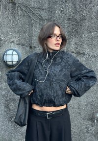 Black zip-up jacket with a floral pattern, oversized sleeves, and an elastic hem, paired with a black belt and black wide-leg trousers.