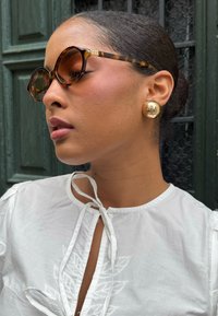 Skildpaddemønstrede solbriller med runde stel, en lys brun tone og guld accents detaljer; store guld cirkulære øreringe; hvid bluse med bindelukning.