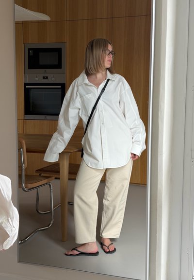 Mujer con el pelo corto de pie descalza frente a un espejo, vistiendo una camisa blanca de gran tamaño, pantalones beige, gafas negras y un bolso cruzado.