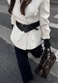 Weißer Blazer mit schwarzem Gürtel, dunkelblaue Culotte-Hose, schwarze Handschuhe und eine braune Handtasche mit goldenen Beschlägen. Strukturierter Stoff sichtbar.