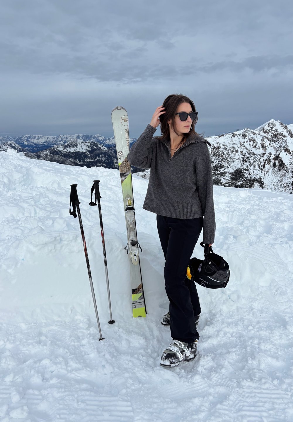 Femme en pull et pantalon foncés debout sur la neige tenant un casque de ski, à côté de skis et bâtons avec des montagnes enneigées en arrière-plan.