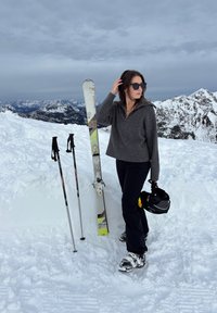 Femme en pull et pantalon foncés debout sur la neige tenant un casque de ski, à côté de skis et bâtons avec des montagnes enneigées en arrière-plan.