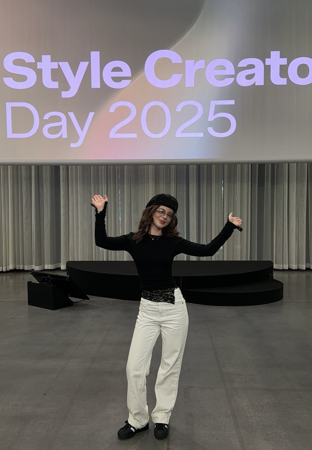Personne portant un haut à manches longues noir, un pantalon blanc et des chaussures noires, avec un béret noir et des lunettes, devant un arrière-plan affichant « Style Creator Day 2025 ».