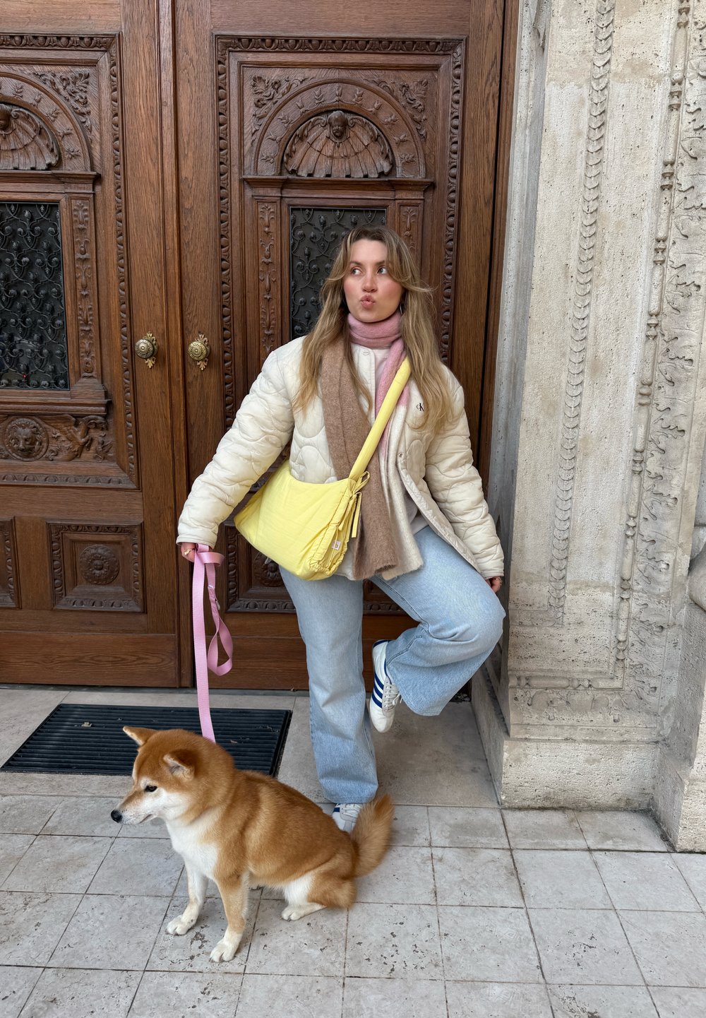 Veste crème matelassée, écharpes rose et marron, sac jaune, jean évasé bleu clair, et baskets blanches, posant avec un chien Shiba Inu.