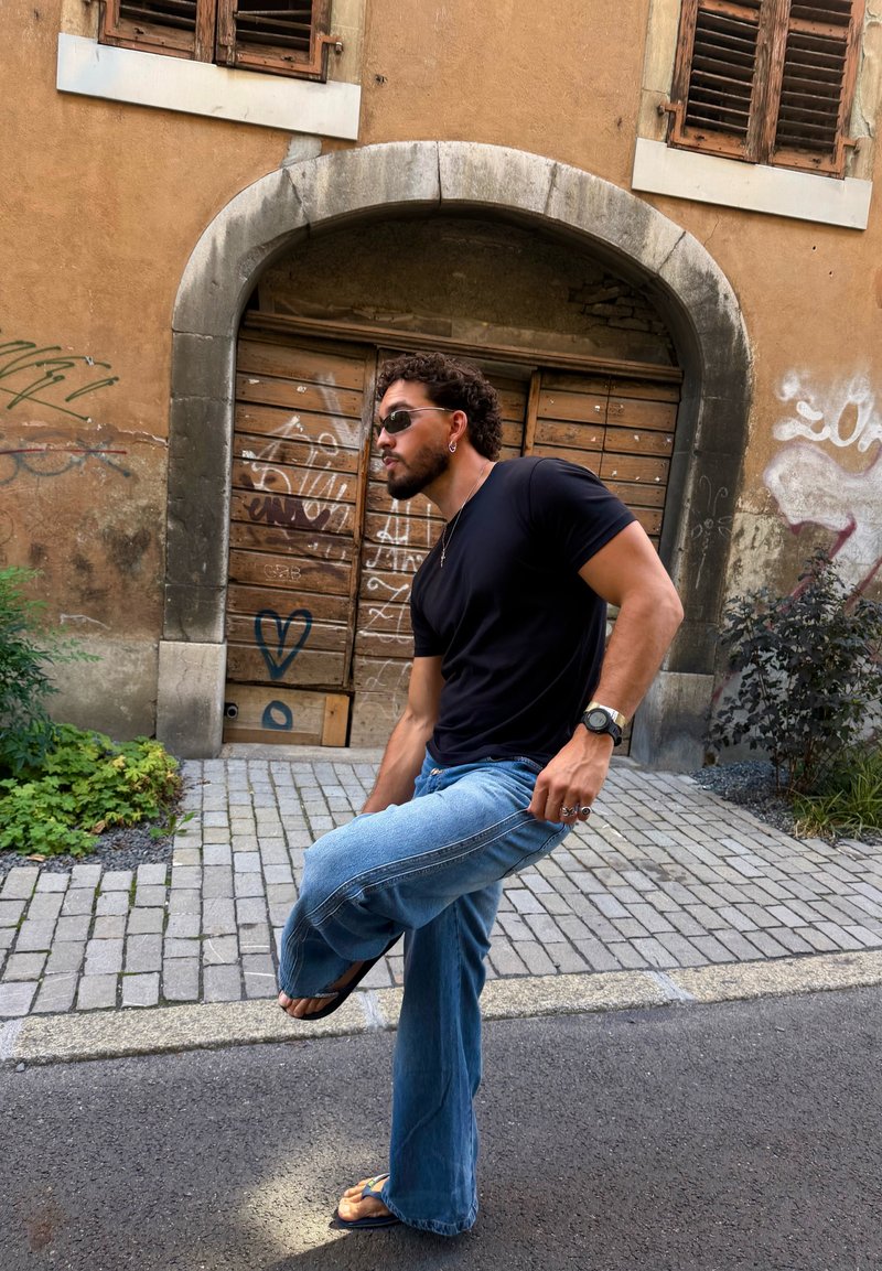 Homme portant une chemise noire, un jean bleu et des sandales, levant une jambe tout en se tenant dans une rue pavée avec une porte en bois couverte de graffiti derrière lui.