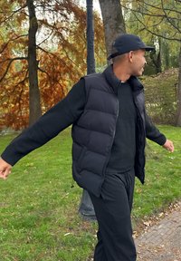 Veste matelassée noire portée sur un pull noir, dotée d'une fermeture éclair et de poches latérales. Portée avec un pantalon noir, sur fond de parc verdoyant.