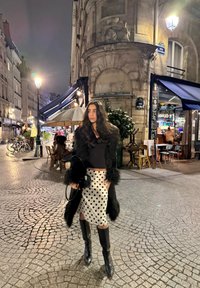 Abrigo de piel sintética negro, top negro ajustado, falda blanca con lunares negros, botas negras hasta la rodilla, de pie en una calle empedrada, ambiente nocturno.