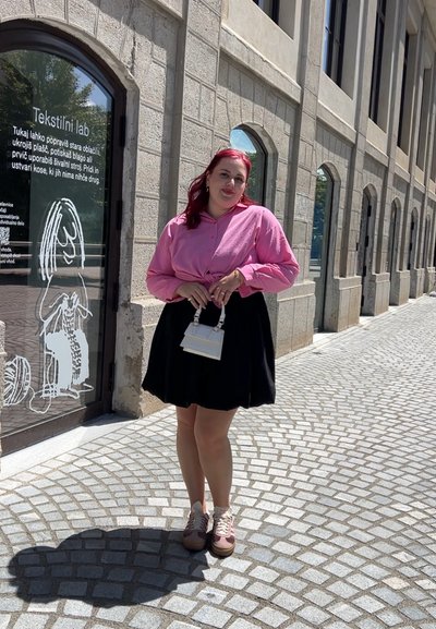 Una mujer se encuentra en una calle de adoquines, vistiendo una camisa de botones rosa y una falda negra, sosteniendo un pequeño bolso blanco. Tiene el cabello rosa.
