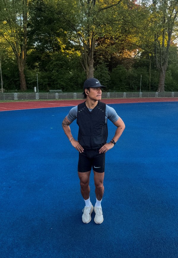 Man in athletic gear with hands on hips stands on blue running track, surrounded by green trees and a fence in the background.