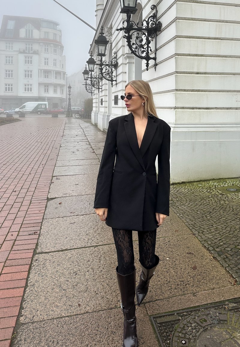 Blazer noir à manches longues avec fermeture à boutons, associé à des leggings en dentelle noire et des bottes en cuir marron montant au genou ; en extérieur dans un décor brumeux.