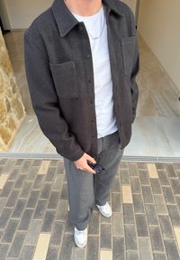 Camisa de botones gris oscuro con textura y dos bolsillos delanteros, usada sobre una camiseta blanca y combinada con pantalones a rayas y zapatillas blancas.