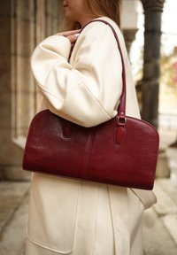 Torbička iz bordo usnja s strukturirano obliko in majhnimi zaobljenimi konci. Ima eno naramnico in subtilne detajle šivov.