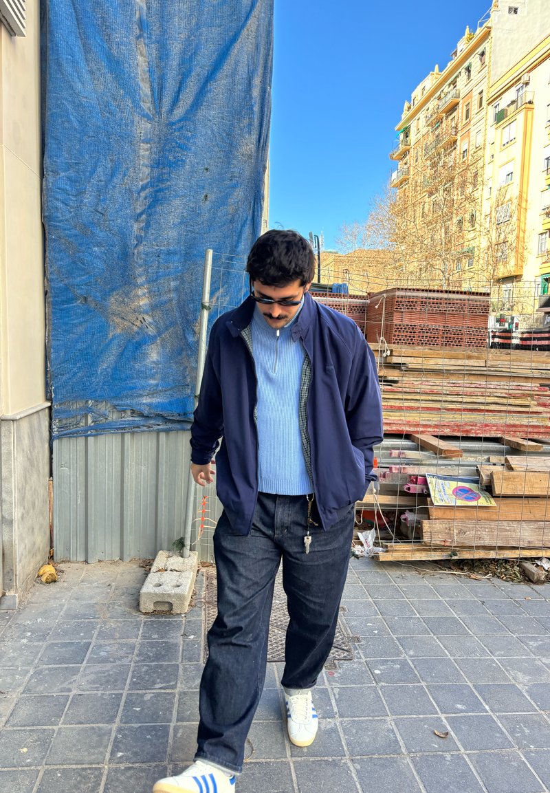 Marineblauwe zip-up jas over een lichtblauw gebreid sweater, donkerblauwe spijkerbroek en witte sneakers met blauwe strepen, wandelend op een bestraat pad.