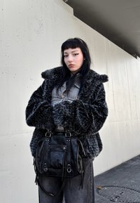 Fekete műszőrme kabát széles mandzsettákkal, alatta kockás ing, fekete bőr táska cipzárokkal és fém részletekkel, világos fal előtt állva.