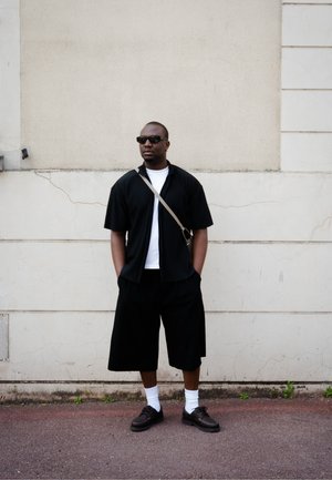 Camisa negra de manga corta con rayas verticales, camiseta blanca debajo, pantalones cortos negros hasta la rodilla, zapatos negros y gafas de sol.