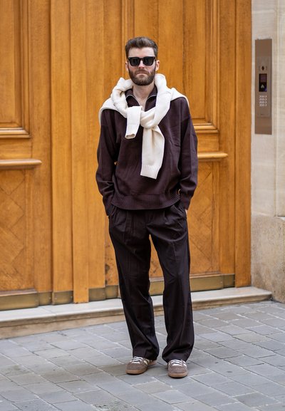 Suéter marrón oscuro con una bufanda crema alrededor del cuello, combinado con pantalones oscuros de corte holgado y zapatillas marrones, sobre un fondo de madera.