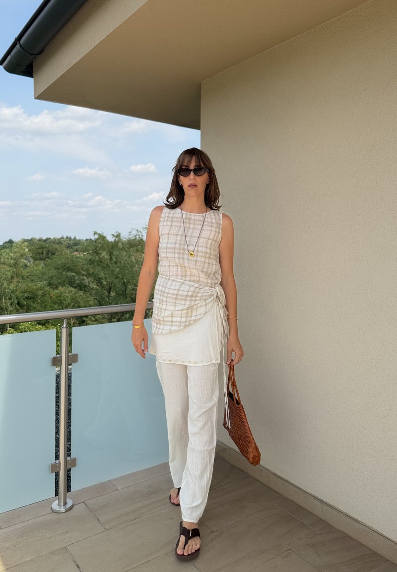 Donna con top a quadri senza maniche, pantaloni bianchi, occhiali da sole e sandali, che tiene una borsa marrone intrecciata su un balcone con alberi sullo sfondo.
