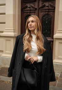 Svart frakk med tynne striper over en hvit topp, holder en svart skinnveske. Langt, bølgete hår og en gullring synlig. Brun dør i bakgrunnen.