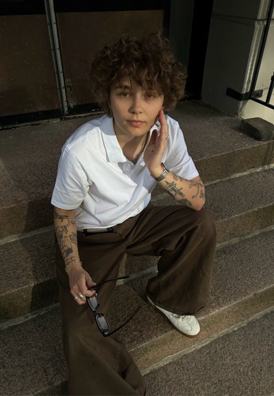 Persona sentada en escalones de piedra, vistiendo una camiseta polo blanca y pantalones anchos marrones. Sostiene gafas de sol, tiene el pelo rizado y tatuajes visibles.