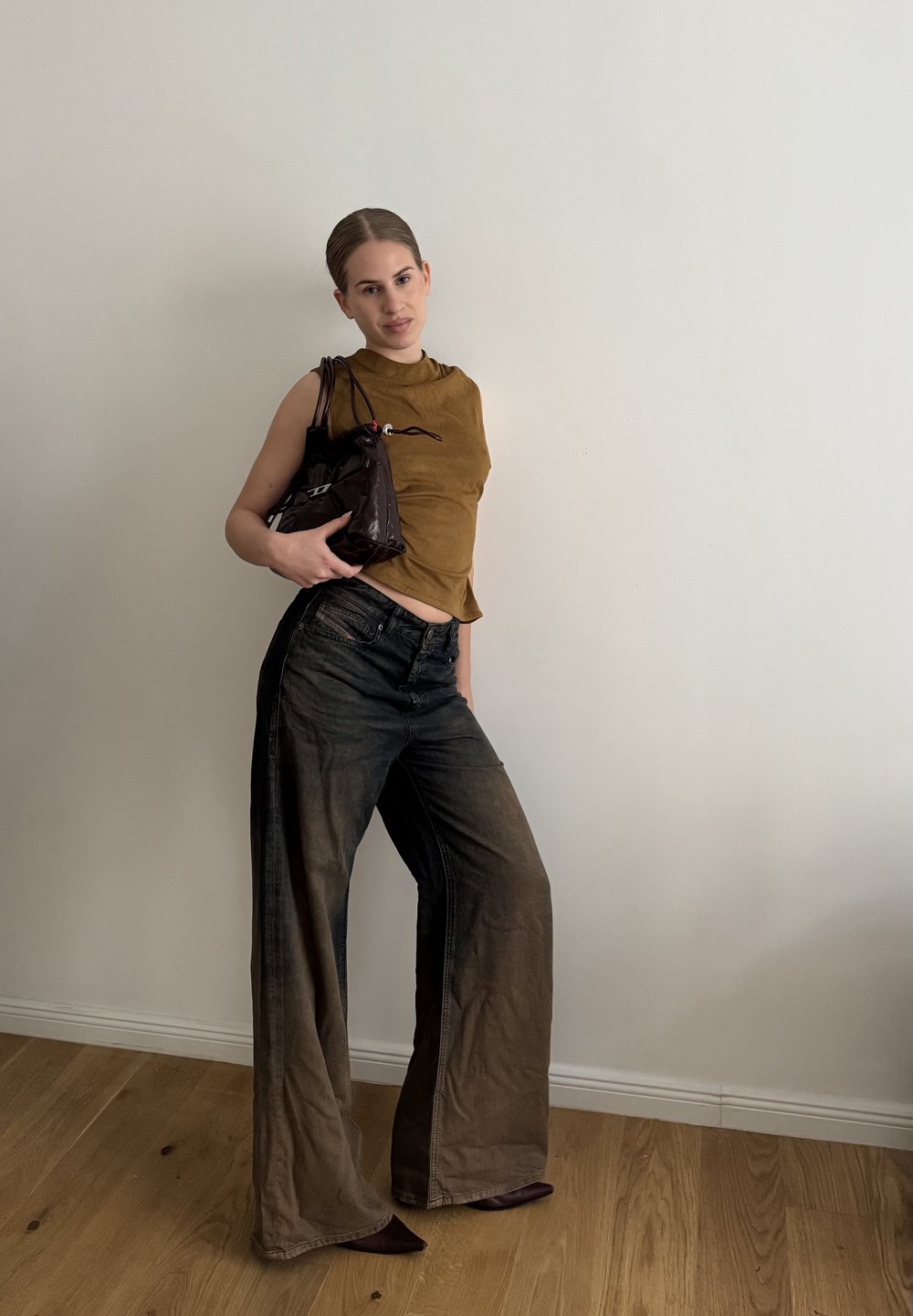 Brązowy krótki bezrękawnik, szerokie wyblakłe czarne dżinsy i błyszcząca czarna torebka. Stylizacja ma gładką fakturę i warstwowy krój.