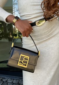 Zwarte patenten leren handtas met een gouden logo, rechthoekige vorm, bovenhandgreep en contrasterende gladde textuur. Draagt met een gouden horloge.