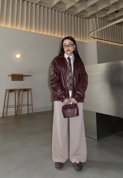 Mujer con gafas, chaqueta de cuero burgundy, corbata de cuadros, pantalones anchos beige, sosteniendo un bolso a juego burgundy en un entorno interior moderno.