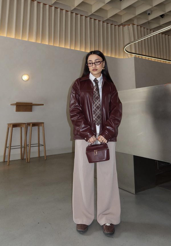 Nő szemüvegben, bordó bőrkabátban, kockás nyakkendővel, bézs színű bő szárú nadrágban, ahogy egy hozzáillő bordó táskát tart modern beltéri környezetben.