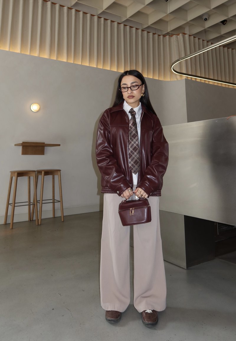 Prillidega naine, burgundi nahktagi, ruuduline lips, beežid laiad püksid, hoiab kaasaegses siseruumis samasugust burgundi käekotti.