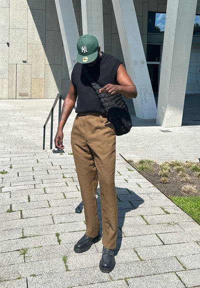 Top de punto sin mangas negro, pantalones de sastre color beige, zapatos negros gruesos y una gorra verde. Bolso de hombro negro texturizado, fondo de piedra gris.