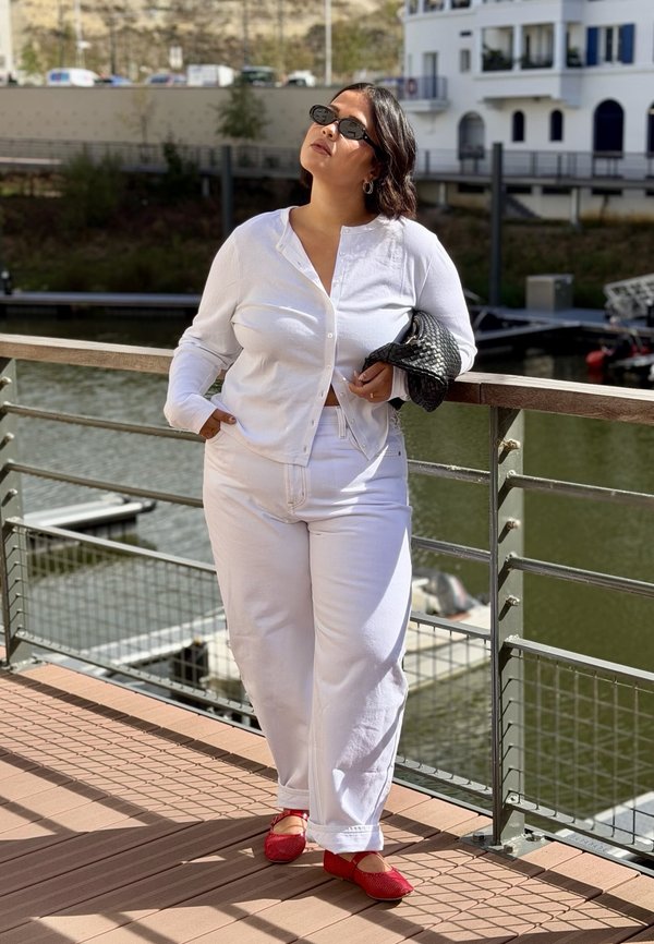 Mujer vestida con ropa blanca y zapatos rojos está de pie en una plataforma de madera junto al agua, sosteniendo un bolso negro tejido y mirando hacia arriba.