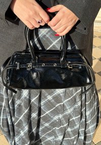 Mains avec du vernis à ongles rouge et une bague tenant un sac à main en cuir noir sur une jupe à carreaux grise et un manteau sombre.