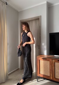 Svart ärmlös topp med vriden detalj, kombinerad med en paljettbeströdd svart kjol. Accessoarerna inkluderar en texturerad svart handväska och platta skor.