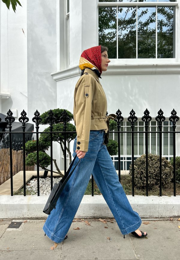 Mujer caminando por la acera con pañuelo rojo y amarillo en la cabeza, chaqueta beige, vaqueros azules anchos, tacones negros y llevando un bolso tote negro.