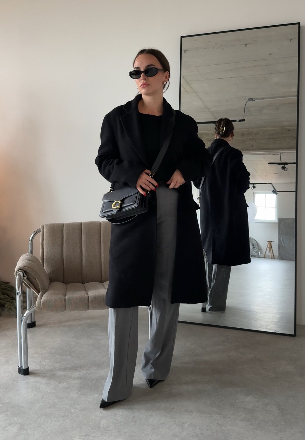 Femme portant un manteau noir, un pantalon gris, des chaussures noires pointues et des lunettes de soleil, debout devant un grand miroir dans une pièce minimaliste.