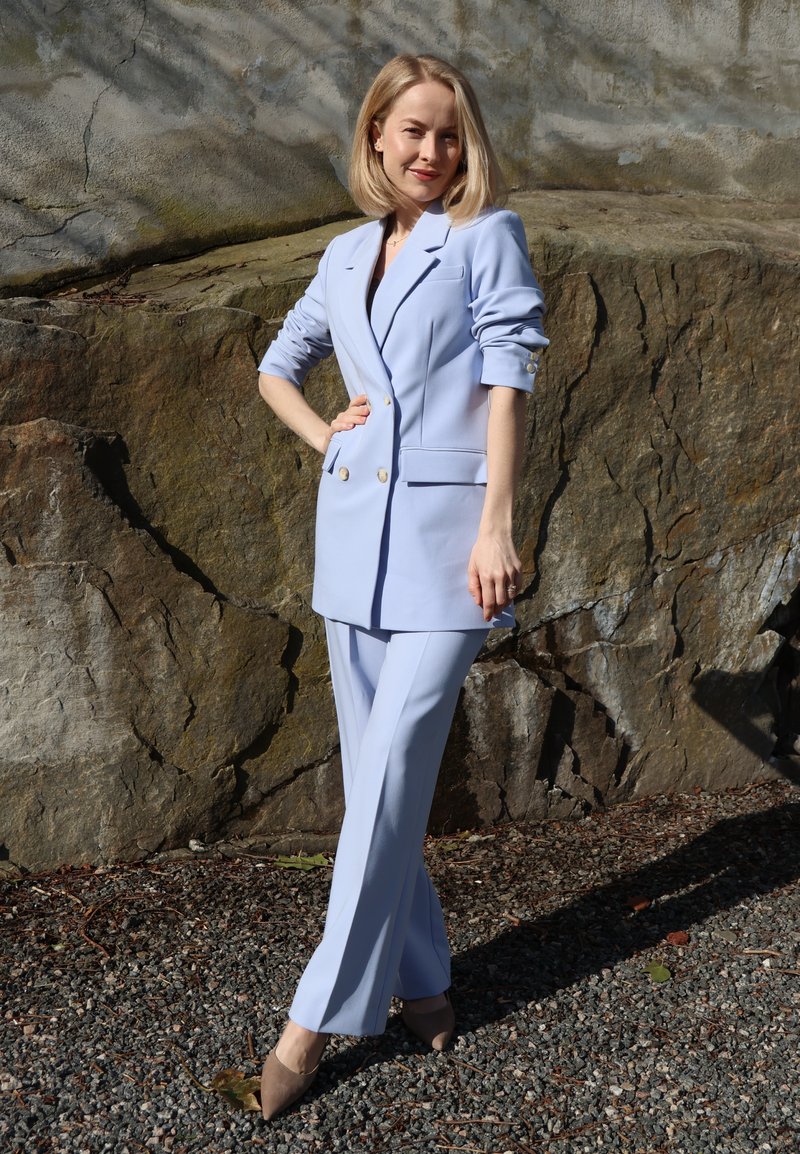 Femme aux cheveux blonds portant un tailleur pantalon bleu clair et des talons beiges, debout sur du gravier devant un grand mur de rochers, souriante, la main sur la hanche.