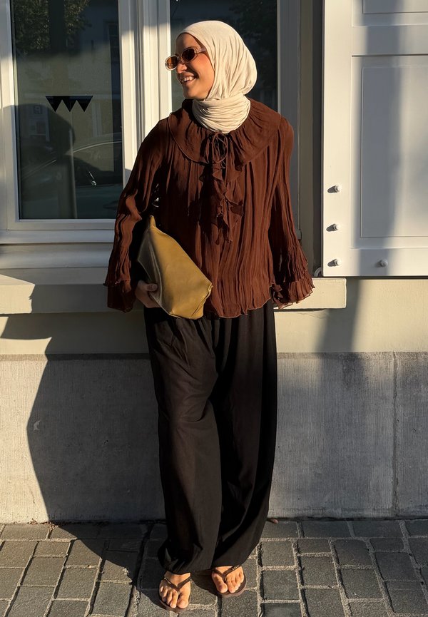 Mujer con pañuelo beige claro en la cabeza, blusa marrón con volantes, pantalones anchos negros, sosteniendo un bolso beige, de pie junto a una ventana en un día soleado.