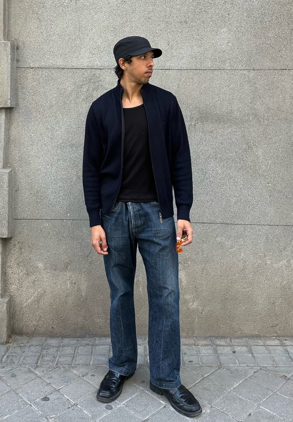Junger Mann mit schwarzer Kappe, schwarzem Reißverschluss-Pullover, schwarzem Hemd und blauer Jeans steht auf einem gepflasterten Gehsteig vor einer grauen Wand.