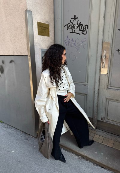 Mujer con cabello largo y rizado junto a la entrada de un edificio, lleva un abrigo blanco, camisa de lunares, pantalones negros y sostiene una gran bolsa tejida.