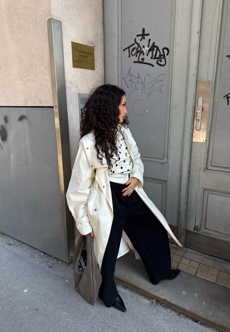 Femme aux longs cheveux bouclés se tient à côté d'une entrée de bâtiment, portant un manteau blanc, une chemise à pois, un pantalon noir, et tenant un grand sac tissé.